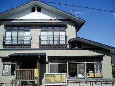 ベージュの外壁と白い切妻屋根の建物で、二階には黒い格子状の手すりが付いた窓が二つ、一階には木製の柵に囲まれた入り口と大きなガラス窓が並び、手前に緑色の掲示板が設置されている戸川児童館外観写真