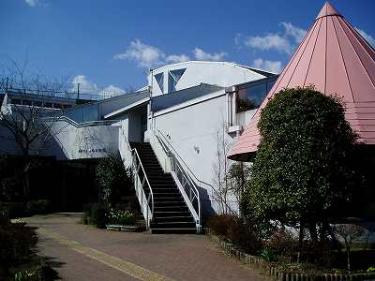 扇型の屋根の白い壁の建物で、階段状に続く構造の中央には黒い階段と白い手すりがあり、右隣にはピンク色の円錐形の屋根を持つ構造物の鶴巻児童室外観写真