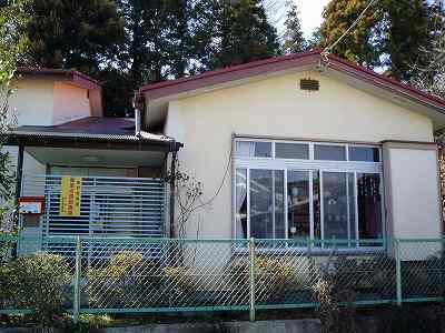 赤い縁取りの屋根を持つ平屋建てのクリーム色の建物で、正面には大きな掃き出し窓と、その左側には緑色の波型フェンスで囲まれた玄関があり、手前には金網フェンスと植栽が配置されている横野児童館外観写真