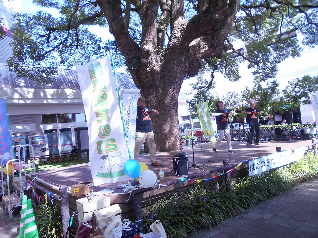 大きな木の前に設置された木で出来たステージにのぼり旗や風船が飾られ、3名の男女が体操をしている写真