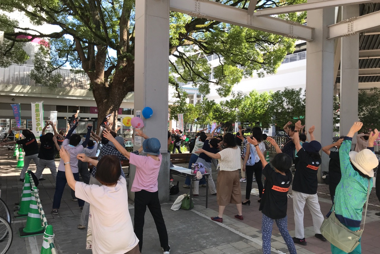 のぼり旗や風船がある広場で多くの人たちが講師の指導に合わせて両手を上に上げ、体操をしている様子の写真