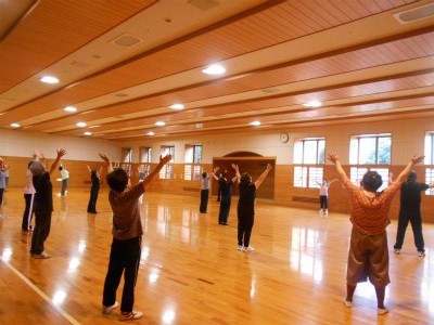 フローリングの広い室内で多くの人たちが両手を上にあげ広げて体操している写真
