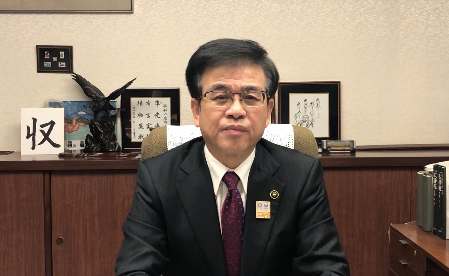 スーツにえんじ色のネクタイを着用した市長が、背景に鳥の彫刻や複数の書、富士山と2羽の折り鶴が描かれた作品が飾られた部屋の椅子に座り、カメラに向かって正面を向いている写真
