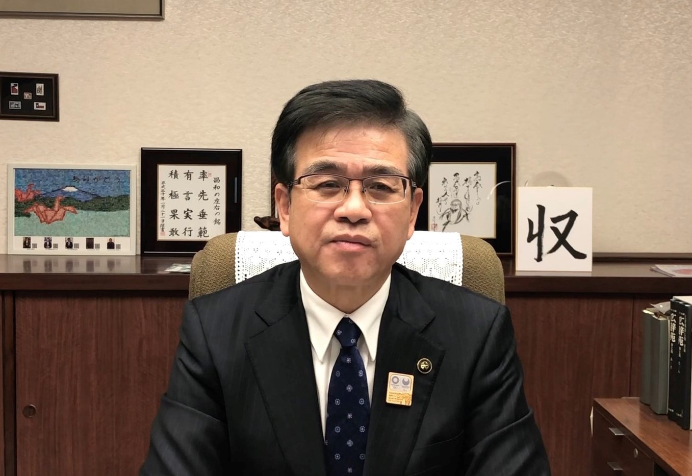 黒いスーツと紺色のネクタイを着用した市長が、富士山と2羽の折り鶴が描かれた作品や額縁に入った書が飾られた部屋の椅子に座り、カメラに向かって真剣な表情でまっすぐ座っている写真（【秦野市民の皆様へ】高橋市長からのメッセージ（令和3年2月3日時点）（YouTube）のサイトへリンク）
