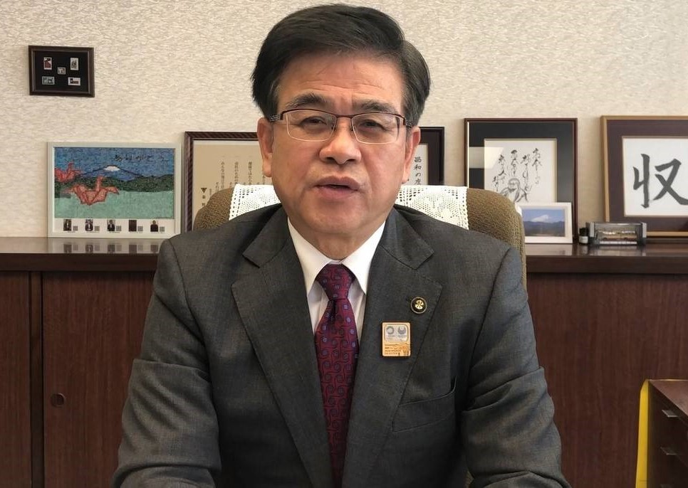 スーツ姿でえんじ色のネクタイをしめた和やかな表情の市長が、富士山と2羽の折り鶴が描かれた作品や富士山の写真、書などが飾られた部屋の椅子にカメラに向かってまっすぐ座っている写真（【秦野市民の皆様へ】高橋市長からのメッセージ（令和3年3月10日時点）（YouTube）のサイトへリンク）