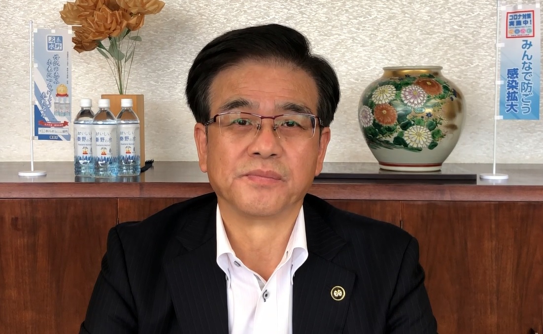 室内でオレンジ色の造花や花柄の花瓶、複数のペットボトル飲料、そして「コロナ対策実施中 みんなで防ごう 感染拡大」と書かれた小さな旗が飾られている木製の棚を背景に、スーツ姿の市長がカメラに向かって座っている写真（【秦野市民の皆様へ】高橋市長からのメッセージ（令和3年5月31日）（YouTube）のサイトへリンク）