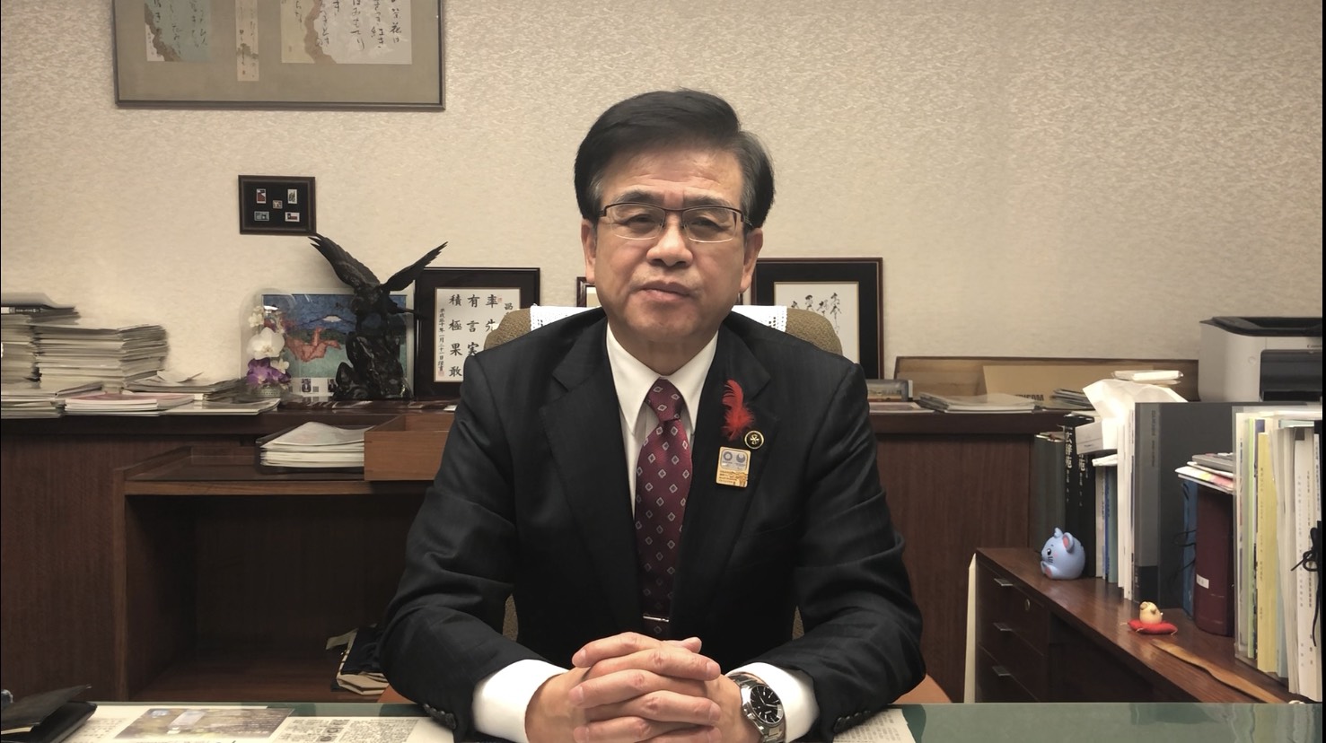 室内で羽を広げた黒い鳥の置物、額に入った書などが置かれている木製の棚を背景に、スーツにえんじ色のネクタイをしめた市長が、デスクに手を置き、正面のカメラを見ている写真（【秦野市民の皆様へ】高橋市長からのメッセージ（令和2年12月4日時点）（YouTube）のサイトへリンク）