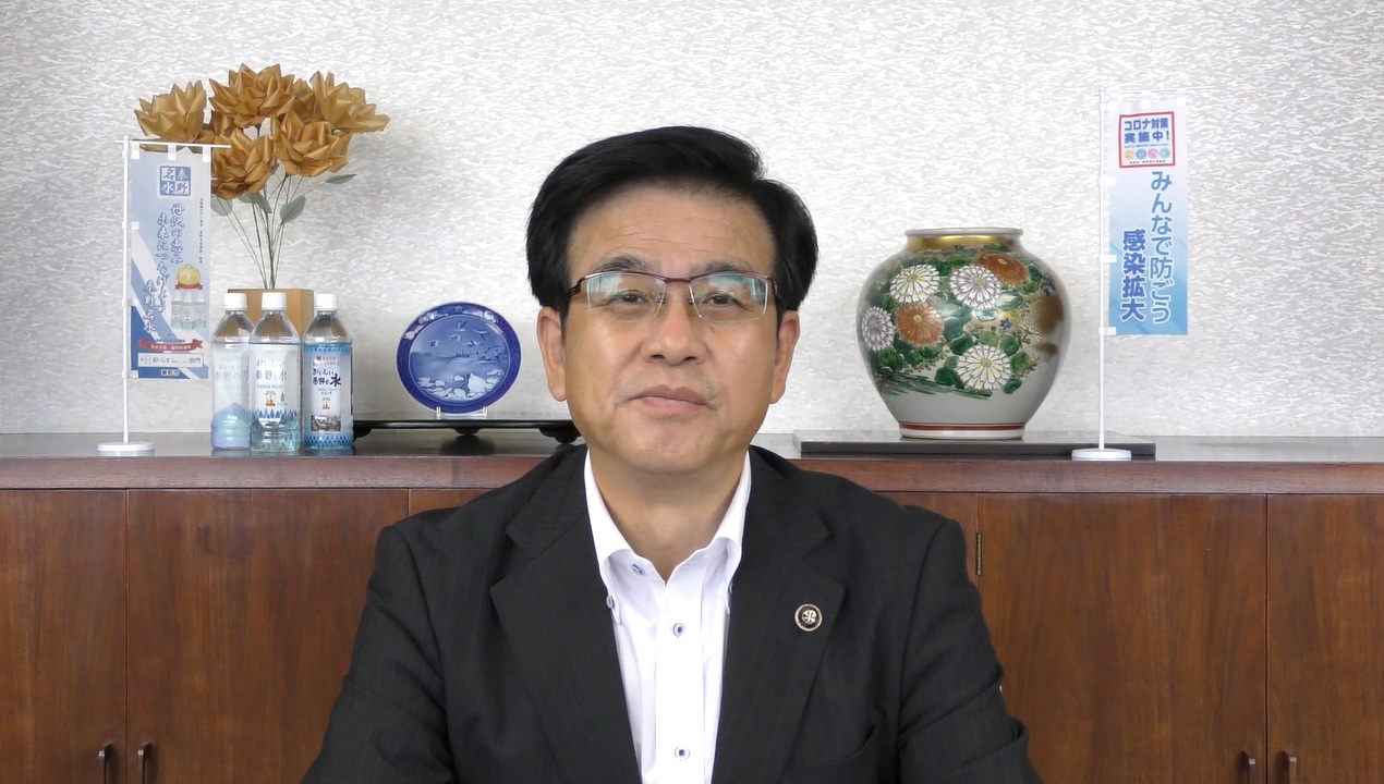 室内で花瓶や皿、ペットボトル飲料、そして「コロナ対策実施中 みんなで防ごう 感染拡大」と書かれた小さな旗が飾られた木製の棚を背景に、白いシャツと黒いジャケットを着た市長が、カメラに向かって正面を向いている様子の写真（【秦野市民の皆様へ】高橋市長からのメッセージ（令和3年7月19日）（YouTube）のサイトへリンク）