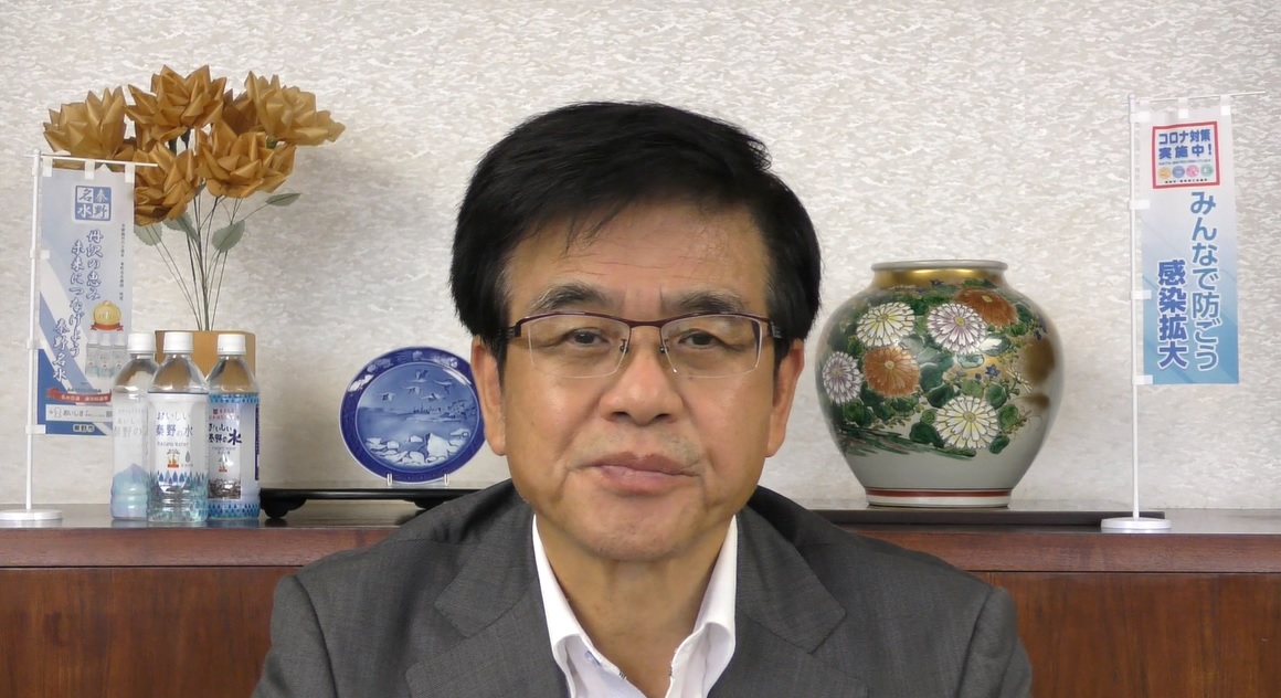 室内で花瓶や皿、ペットボトル飲料、そして「コロナ対策実施中」と書かれた小さな旗が飾られた棚を背景に、白いシャツにグレーのジャケットを着た市長が、カメラに向かって正面を向いている様子の写真（【秦野市民の皆様へ】高橋市長からのメッセージ（令和3年7月30日）（YouTube）のサイトへリンク）