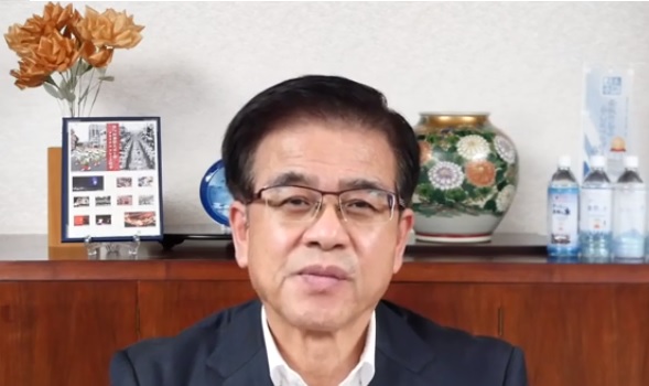 室内で花やペットボトル飲料などが置かれた木製棚を背景に、白いシャツにジャケットを着た市長が、カメラに向かって座っている様子の写真（【秦野市民の皆様へ】高橋市長からのメッセージ（令和3年9月30日）（YouTube）のサイトへリンク）