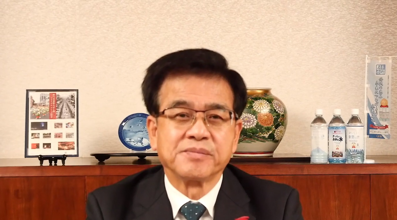 室内で花柄の花瓶や皿などの美術品、そして複数のペットボトル飲料が飾られた木製棚を背景に、スーツを着た市長がカメラに向かって正面を向いている様子の写真（【秦野市民の皆様へ】高橋市長からのメッセージ（令和3年10月22日時点）（YouTube）のサイトへリンク）