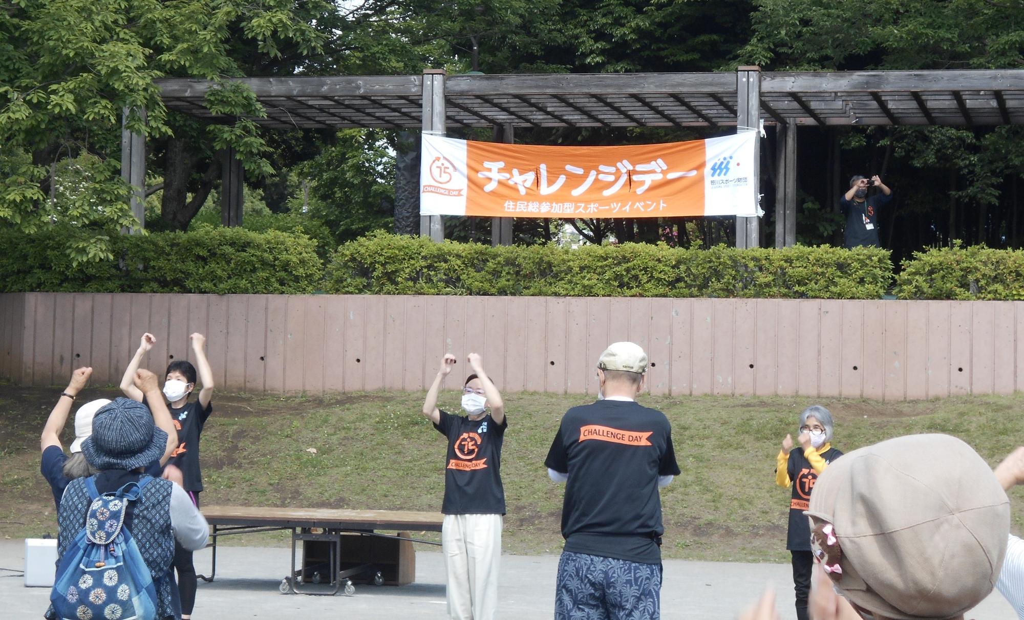 広場の高台にあるパーゴラの柱に「チャレンジデー」と描かれたオレンジ色の横断幕がくくられ、広場ではたくさんの人たちが両手で握り拳を作った状態で上にあげ、体操をしている様子の写真