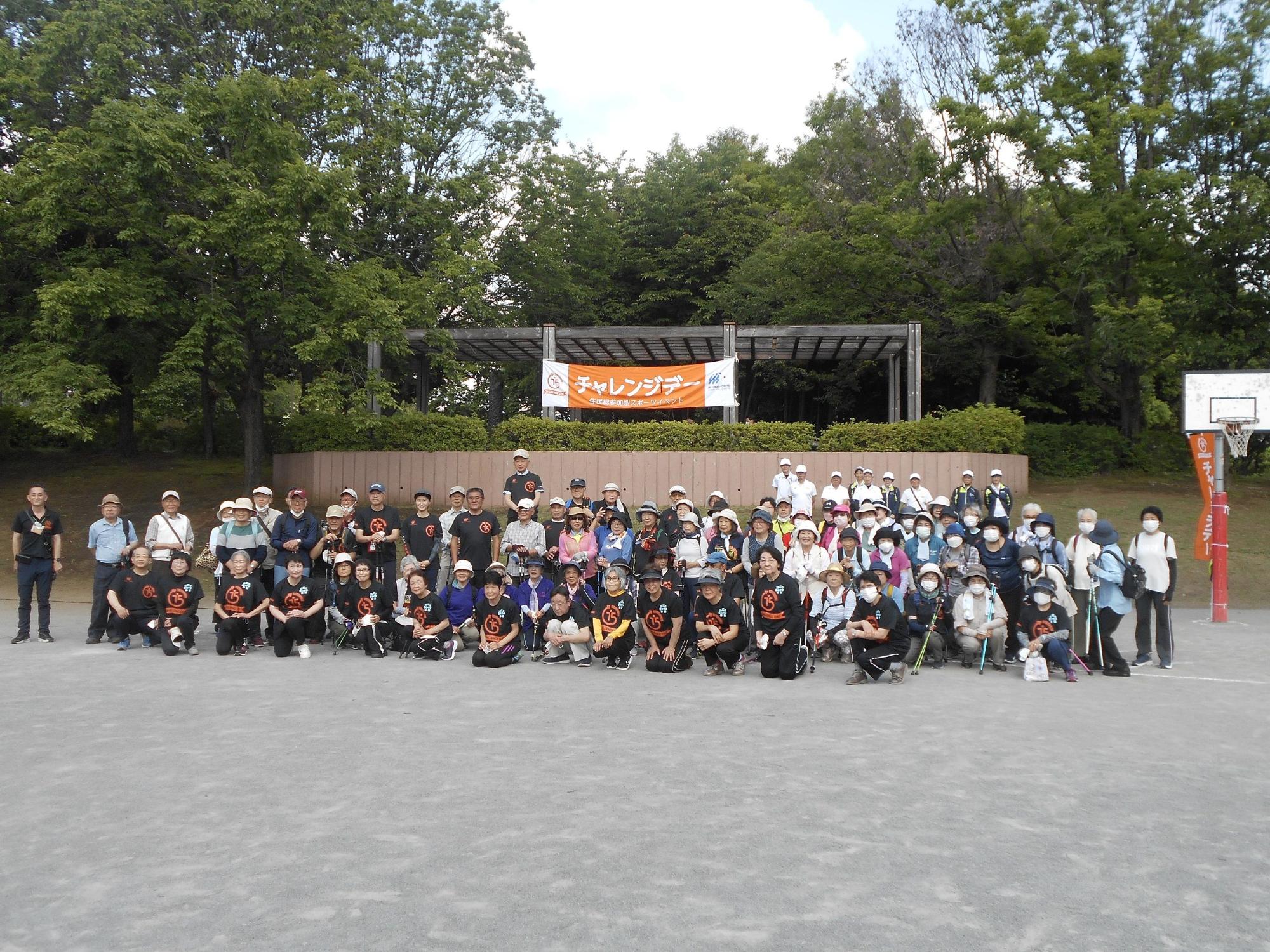 広場の高台にあるパーゴラの柱に「チャレンジデー」と描かれたオレンジ色の横断幕がくくられ、広場では前列でしゃがんでいる人々と後列で立っている人々が記念撮影をしている様子の写真
