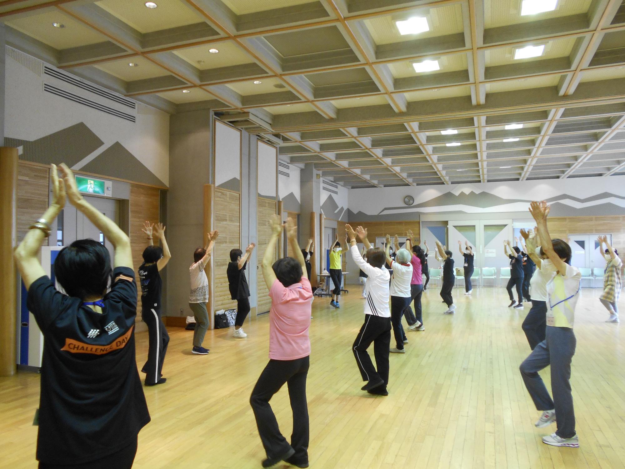 フローリングの広い室内で、たくさんの高齢者が講師の指導に合わせて手のひらを合わせた状態で頭の上に上げ、体操をしている様子の写真