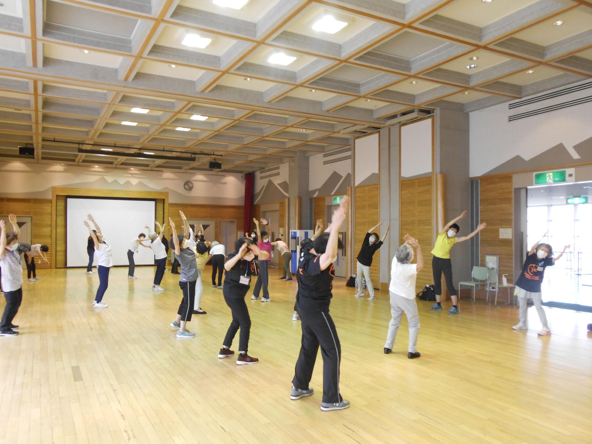 フローリングの広い室内で、たくさんの人々がマスクを着用し、両手を上に上げて体操をしている様子の写真