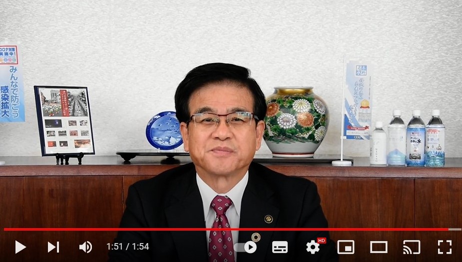 室内で花瓶や水のボトルなどが飾られた棚を背景に、黒いスーツと赤いネクタイを着用した市長が、笑顔で座っている動画再生画面の画像（高橋市長からのメッセージ（令和4年3月22日）（YouTube）のサイトへリンク）
