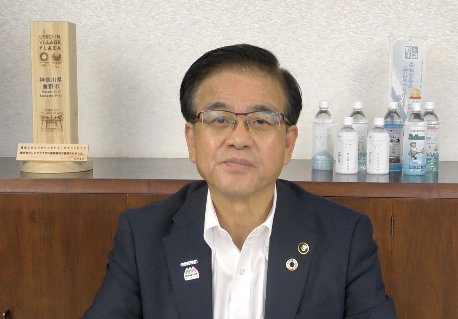 室内で複数のペットボトルや記念品が置かれた木製の棚を背景に、濃紺のジャケットと白いシャツを着た市長が、カメラをまっすぐ見つめている上半身の写真（【秦野市民の皆様へ】高橋市長からのメッセージ（令和4年8月3日）（YouTube）のサイトへリンク）