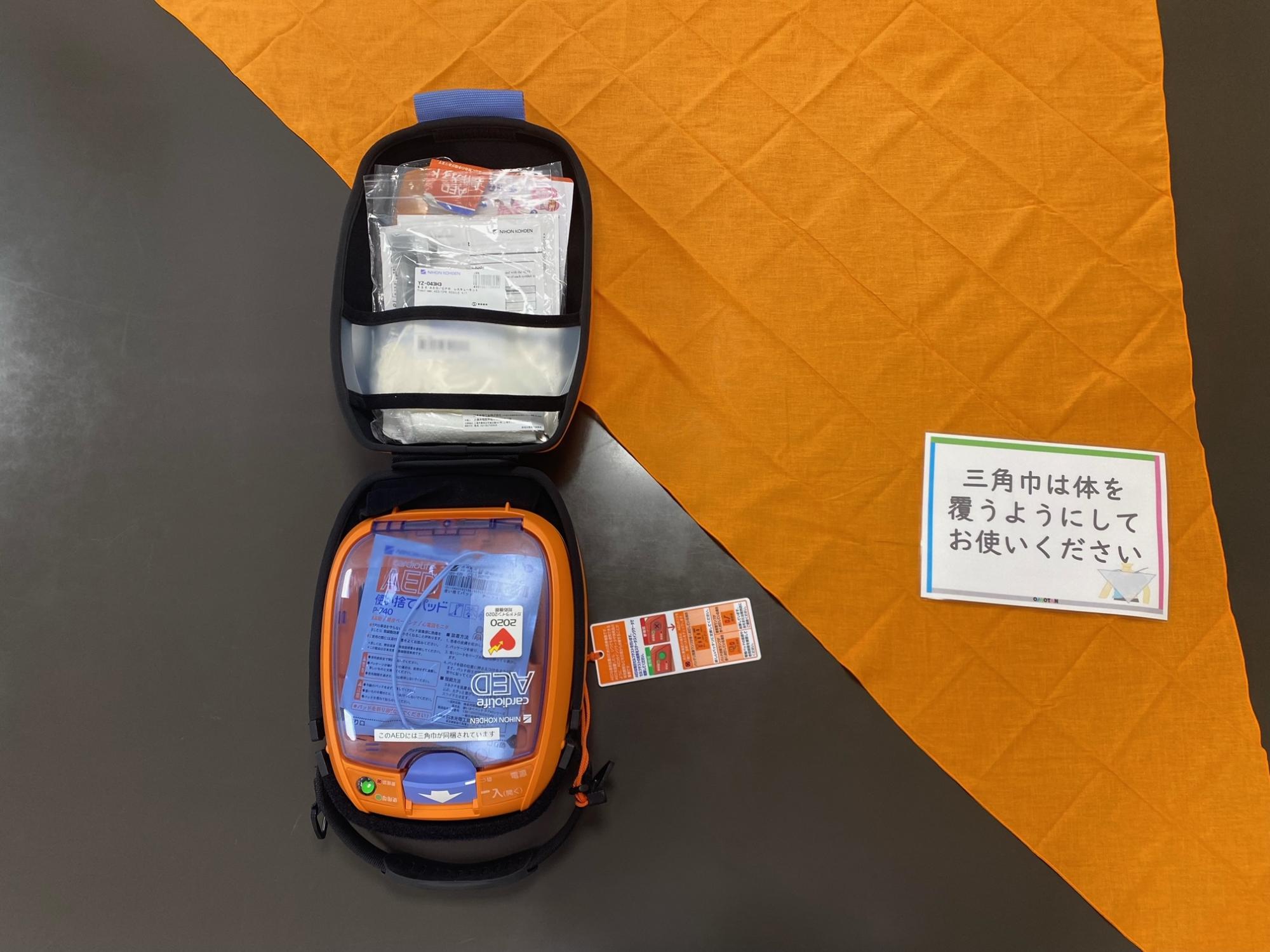 ケースが開かれた状態のAEDが置かれ、その右側にはオレンジ色の三角巾が広げられ、「三角巾は体を覆うようにお使いください」と書かれたカードが添えられている写真