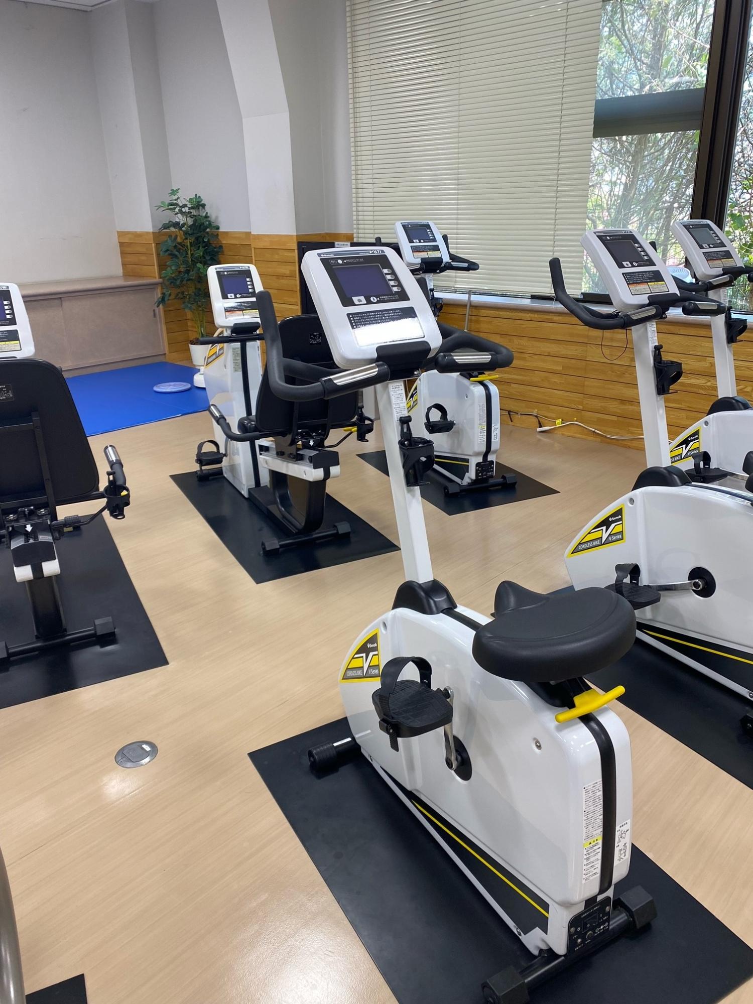自然光が入る大きな窓に面したトレーニングルームに、白と黄色のリカンベントバイク型のフィットネスバイクが等間隔で複数台設置されている写真
