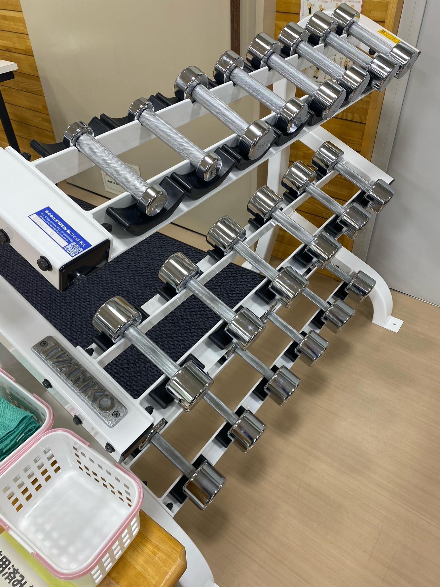 木目調の床とクリーム色の壁のトレーニングルームに、白く傾斜した専用ラックに、光沢のあるクロムメッキ加工が施された、様々な重さのダンベルが整然と段違いで置かれている写真