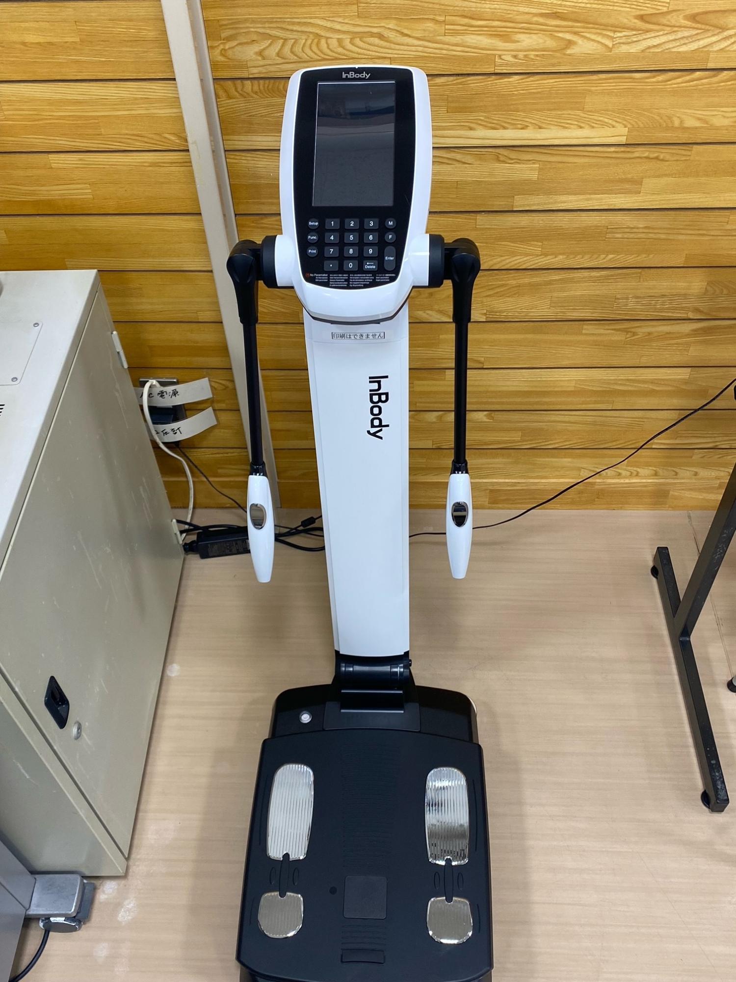 白色で細長いタッチスクリーン付きの本体と黒い台座、両脇に握り手がある体組成分析装置が正面から写された写真