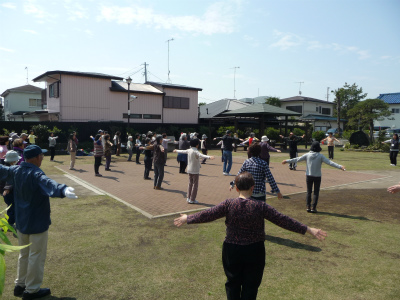 住宅街の公園で多くの人々が円形に広がり、両腕を伸ばして体操をしている様子の写真