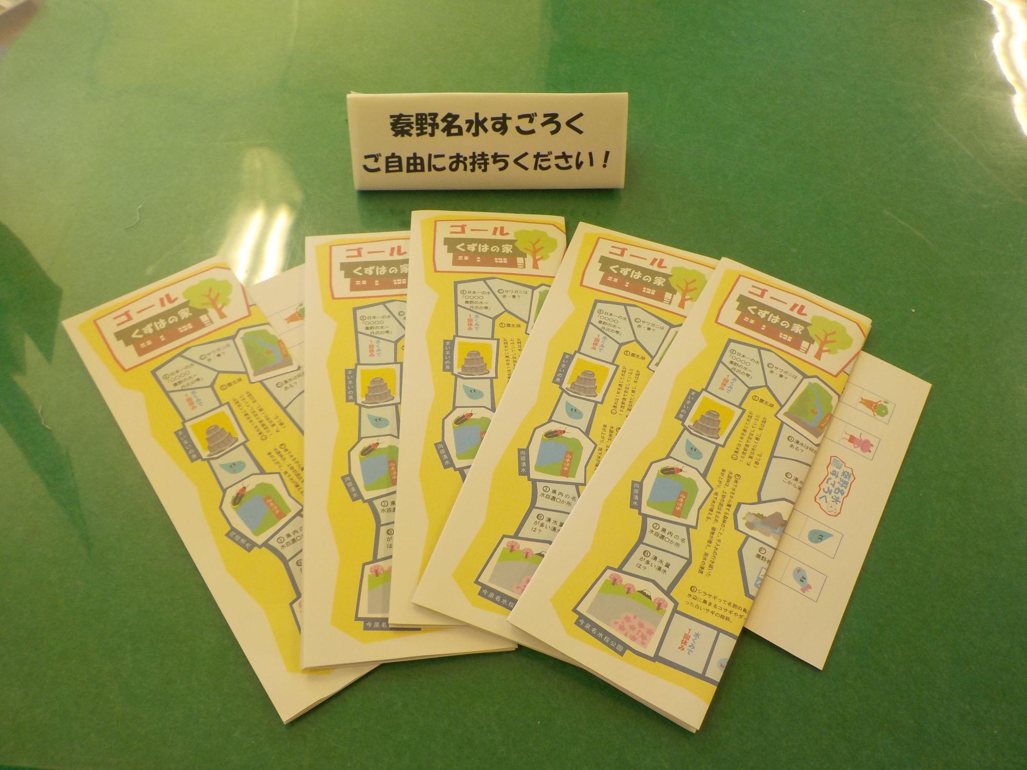 「秦野名水すごろく ご自由にお持ちください！」と書かれた札の手前に折った状態の5枚のすごろくが扇状に並べられている写真