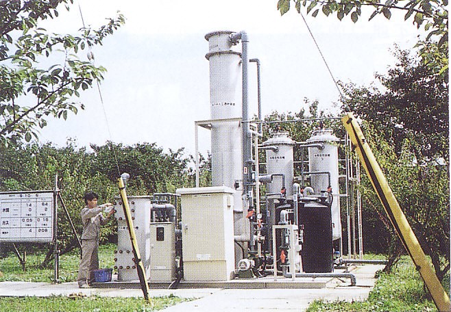 木々に囲まれた敷地内に大きな塔やタンク、配管が複雑に組み合わさった地下水浄化装置が設置されており、作業服姿の男性作業員が機器の操作をしている様子の写真