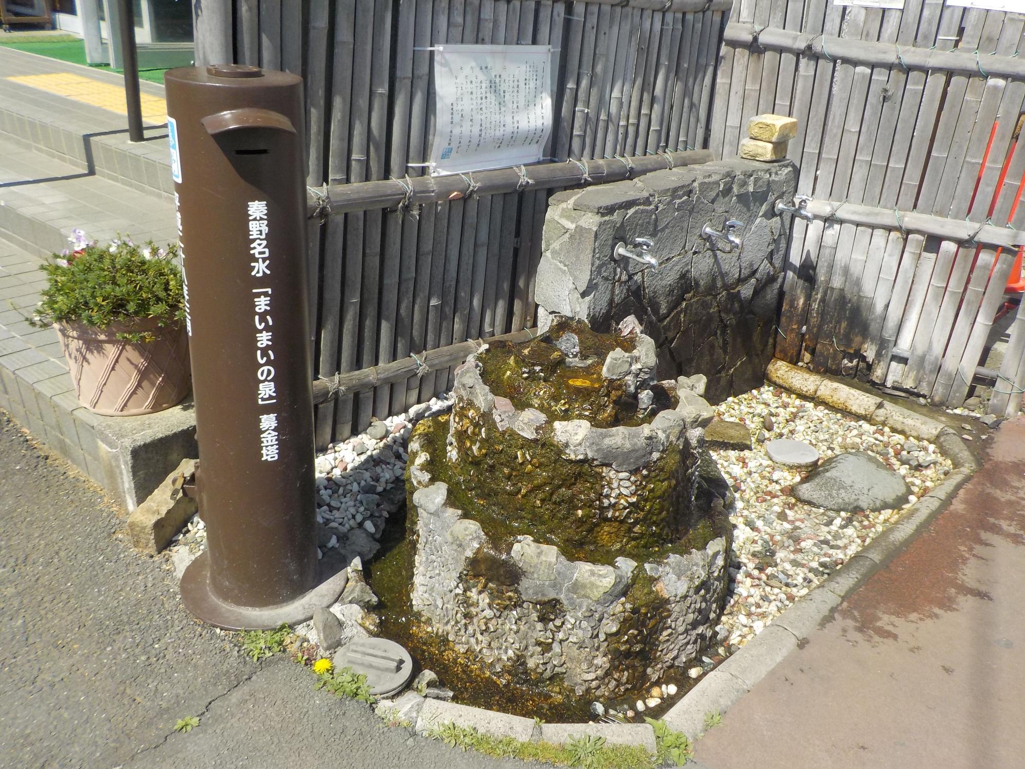 背景の石壁には複数の蛇口が設置されている手前に、茶色の円柱型の表面に「秦野名水「まいまいの泉」募金箱」と書かれた募金箱と、傍に水が溢れ出ている石組みの湧水施設が写っている写真