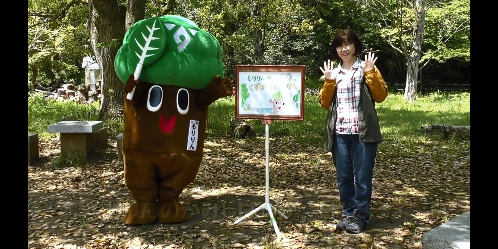 豊かな緑の木々の下に立つ、木の幹のような茶色の胴体と大きな緑の頭に白い葉の模様がある着ぐるみのキャラクター「もりりん」が、両手を広げて笑顔の女性と並び、中央に紙芝居が立てられている写真