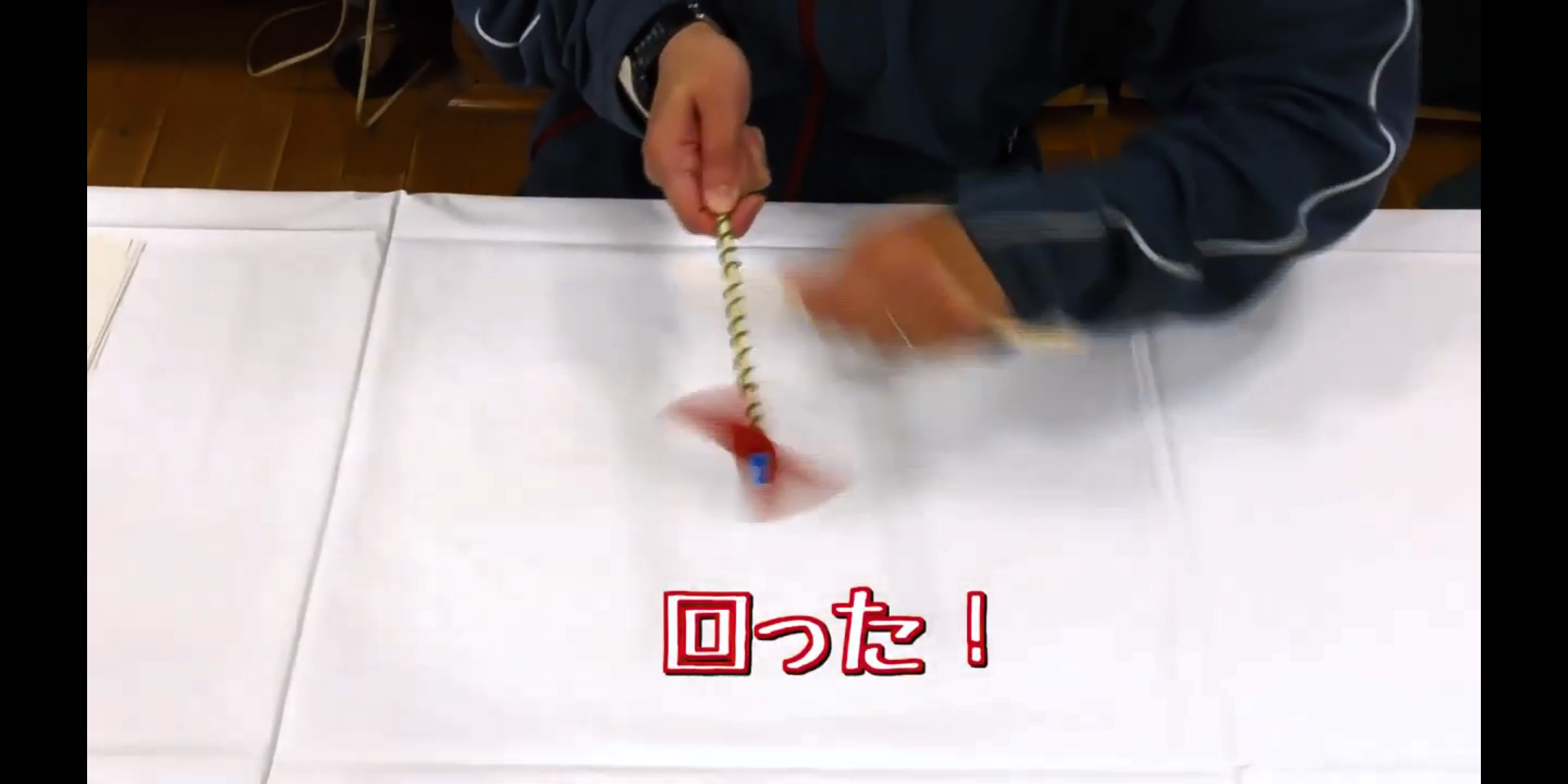 中央下に「回った！」と記載され、紺色の長袖服を着た人物が、白いテーブルの上に置いてある、黄色と緑色の棒の先についた赤と青の羽が勢いよく回転している竹とんぼを両手で持って遊んでいる様子の写真