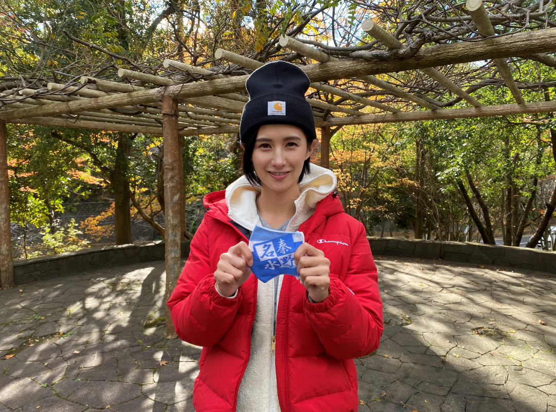 木々に囲まれた屋外で、胸元で「名秦野水」と書かれた青い布のようなものを両手で持っている、黒いニット帽と赤色のダウンジャケットを着た加藤優さんによる啓発動画のYouTube画面が表示されたスクリーンショット画像