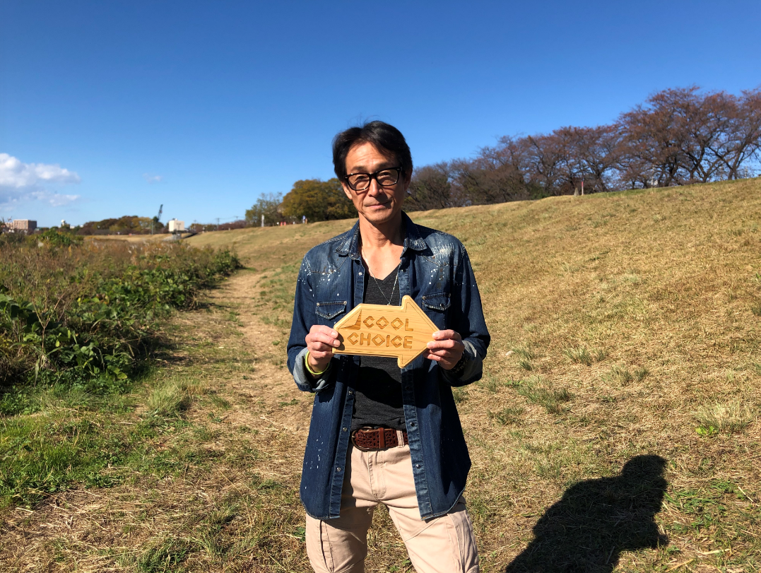 木々が立ち並んだ河川敷に立ち「COOL CHOICE」と刻まれた木製の矢印型パネルを両手で持った、吉田栄作さんによる啓発動画のYouTube画面が表示されたスクリーンショット画像