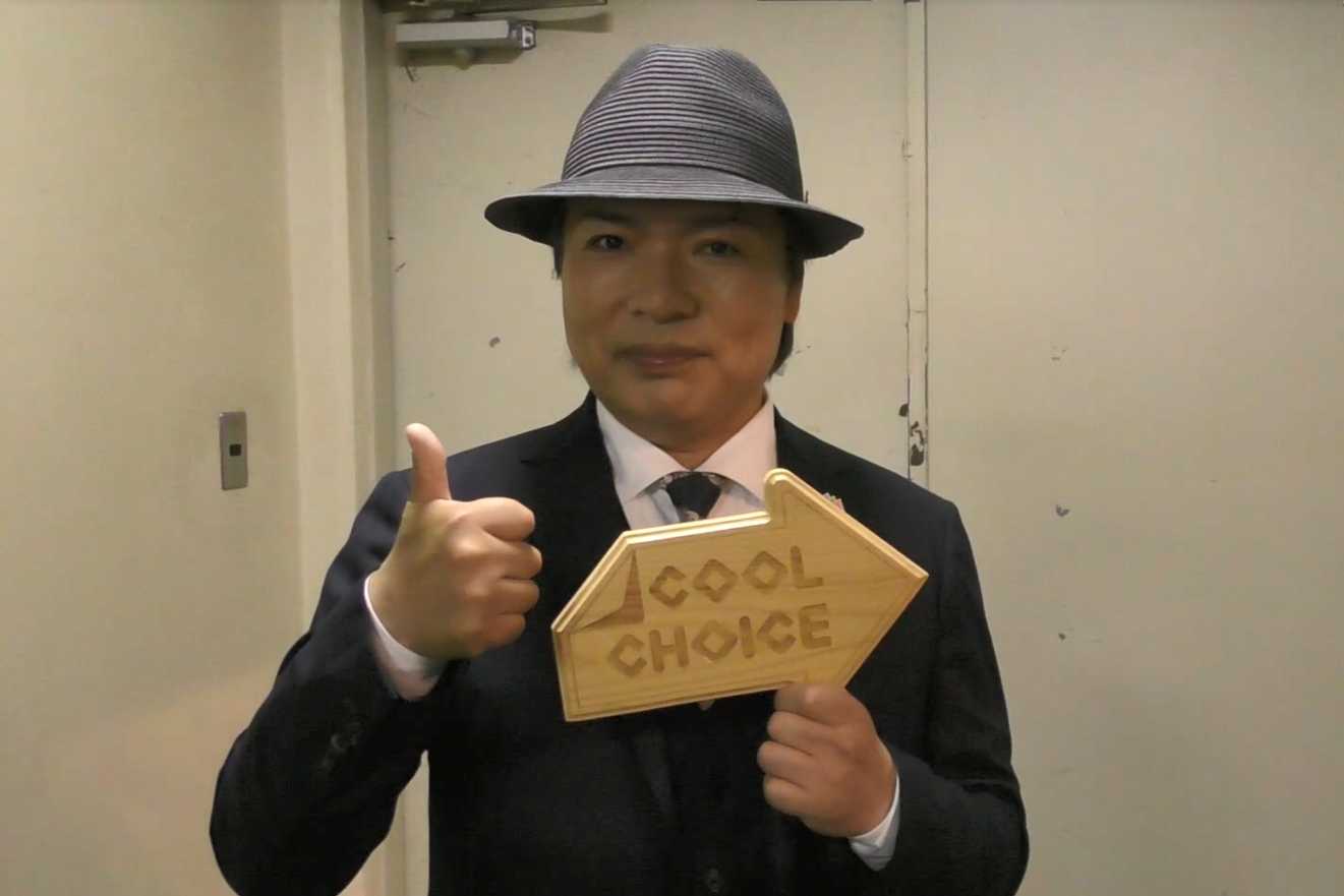 屋内の白い扉の前に立ち、「COOL CHOICE」と刻まれた木製の矢印型パネルを右手で持つ左手でサムズアップポーズをとった、黒いスーツにグレー色の帽子を被ったルナシー真矢さんによる啓発動画のYouTube画面が表示されたスクリーンショット画像