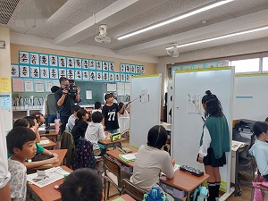 教室で生徒がホワイトボードを使って発表している様子を、カメラマンが撮影しており、教師や他の生徒たちが静かに見守っている、公開授業の写真