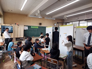 複数の生徒がホワイトボードに向かって発表しており、教室内で他の生徒や教師がその様子を見ている、公開授業の写真