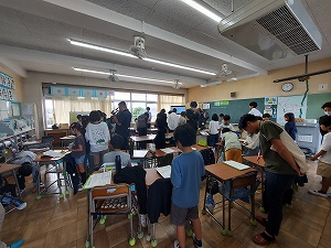 教室内で多くの生徒が机に座ったり立ったりして活動しており、教育の一環としての発表や交流が行われている様子を写した、公開授業の写真
