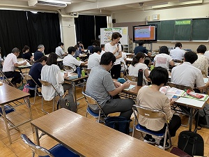 教室で複数の人が机に座り、前方のスクリーンとホワイトボードを見ながら講義を受けている様子を写した、指導者養成講座の写真