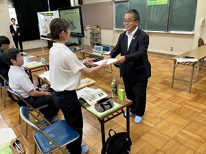 教室で一人の参加者が前に立ち、もう一人から書類を受け取っている場面を写した、任命書が授与の様子の写真