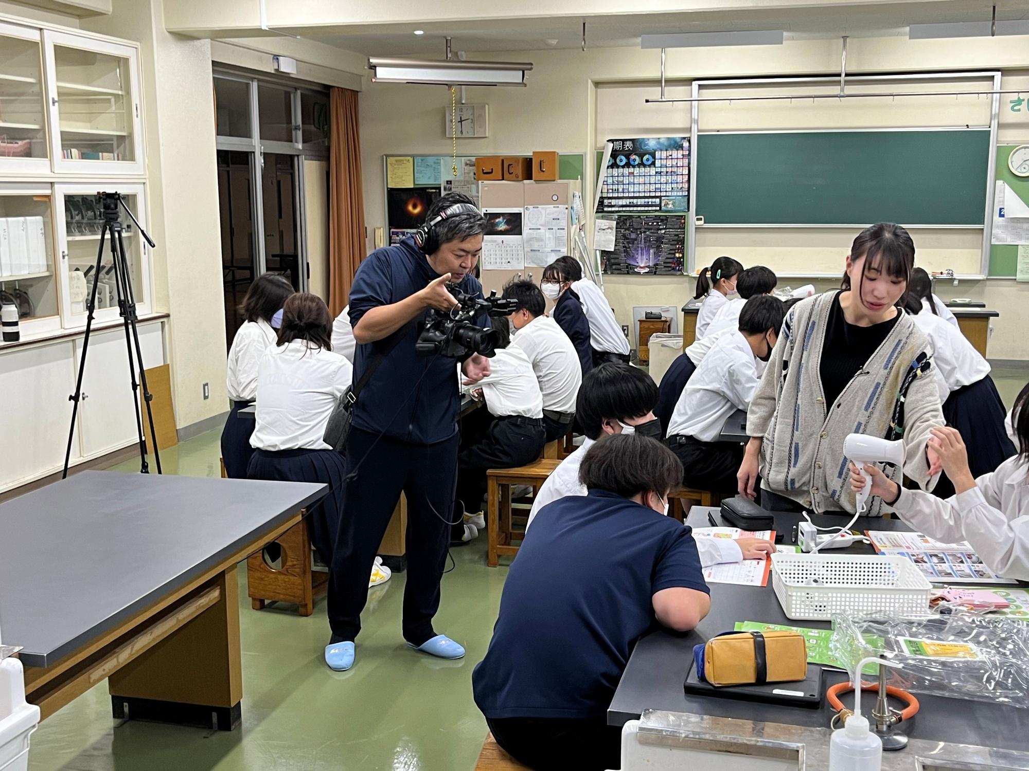 教室で生徒たちが実験を行っている様子をカメラマンが撮影しており、省エネ学習の一環として活動が記録されている、授業撮影会の写真