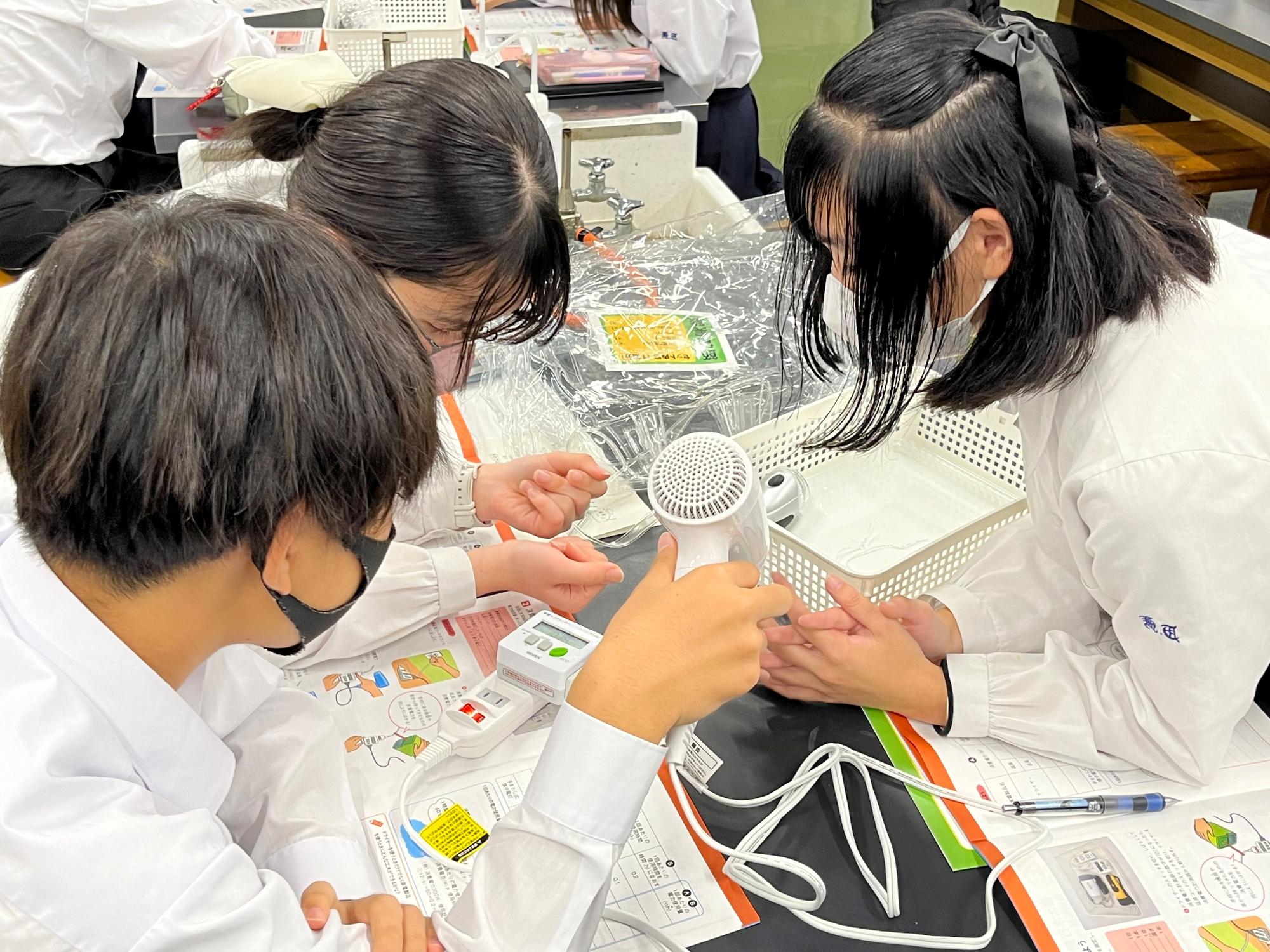 資料が置かれた机の上で、生徒たちがドライヤーを使って電力消費に関する実験を行っている様子を写した、授業撮影会の写真