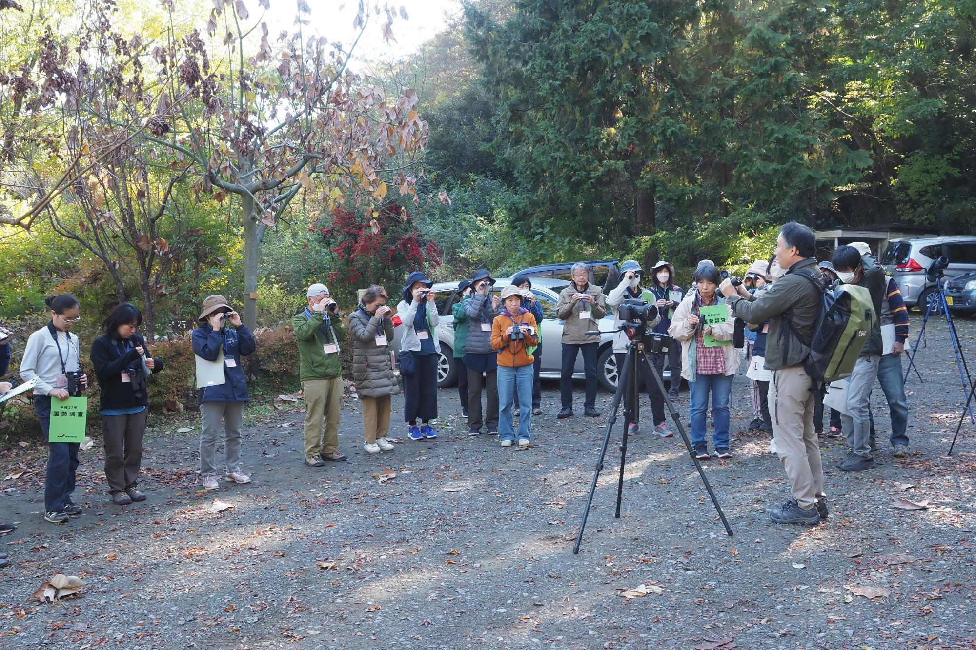 カメラや双眼鏡を持った人たちが、秋の木々が茂る屋外の砂利の広場に集まり、三脚に固定された望遠鏡のようなものを操作する講師らしき男性の説明を聞いている講座の様子の写真