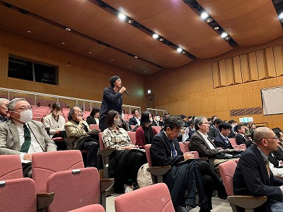 他の観客が聞いているなか、観客席の後方にマイクを持っているニット帽を被った男性が質問を行っている様子の写真