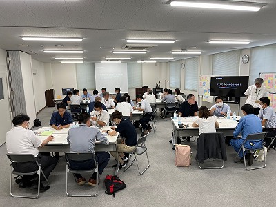 奥の壁にはプロジェクターと大型モニターにプレゼンテーション資料が映し出されている会議室で、白とグレーの机を囲みカジュアルな服装を含むビジネスウェア姿の男女が着席してディスカッションを行っている様子の写真