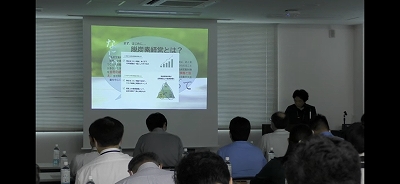 スクリーンに「脱酸素経営とは？」というタイトルとグラフや図を含む資料が投影され、部屋の右奥で講師が受講者に対して説明を行っている様子を後方から捉えた写真
