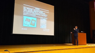 ステージ上に資料が映し出されたスクリーンが設置され、檀上の横に立つスーツ姿の男性がマイクを持ちながら話をしている様子の写真