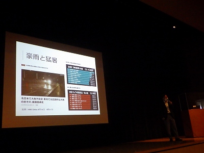 暗い会場で、スーツ姿の講師が右側に立ち、スクリーンには「豪雨と猛暑」というテーマのもと、雨が降っている夜に走行する車の写真と、青いグラフで3時間の降水量、赤いグラフで最高気温を示したニュース記事の画像が投影されている第一部のセミナーの様子の写真
