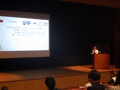 照明の落ちた会場のステージ上で、木製の演台に立つ男性講師が聴衆に向かって話しており、左側の大きなスクリーンには「はじめに」というタイトルの下で「二酸化炭素の排出拡大」に関する文字情報とロゴが提示されている第二部のセミナーの様子の写真