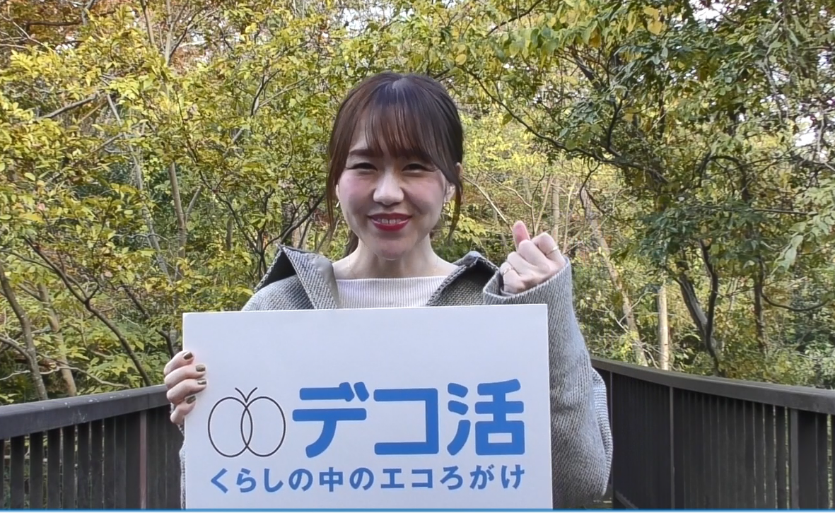 木々が立ち並ぶ茶色の柵の前に立ち、「デコ活くらしの中のエコろがけ」と書かれたボードを持ち、左手でガッツポーズをした、立石純子さんのYouTube画面が表示されたスクリーンショット画像