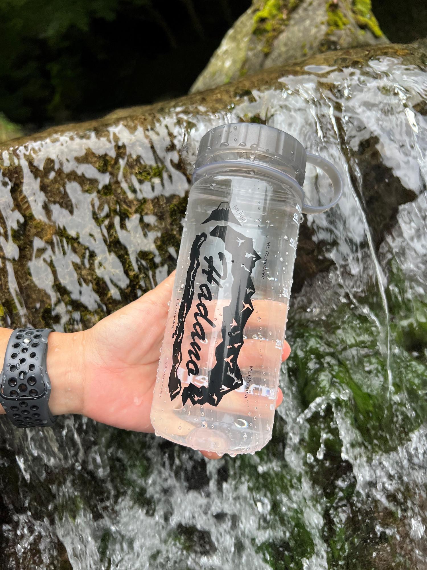 水が流れている大きな岩をバックに、黒い腕時計をした手で持った、アルファベットで秦野と書かれたクリアボトルを掲げている写真